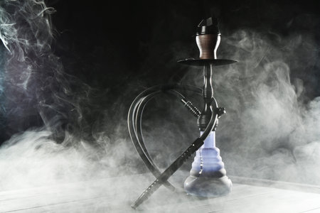 Hookah on wooden table with smoke against black background