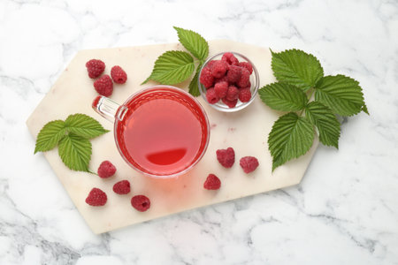 Aromatic raspberry tea in glass cup, berries and green leaves on white marble table, top view