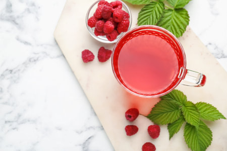 Aromatic raspberry tea in glass cup, berries and green leaves on white marble table, top view. Space...
