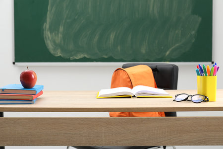 Different school stationery, glasses and red apple on wooden table near blackboard indoors