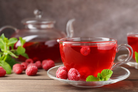 Tasty raspberry tea in glass cup, teapot, berries and mint on wooden table, closeup