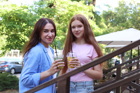 Portrait of beautiful mother and her teenage daughter with refreshing drinks on stairs outdoorsの写真素材