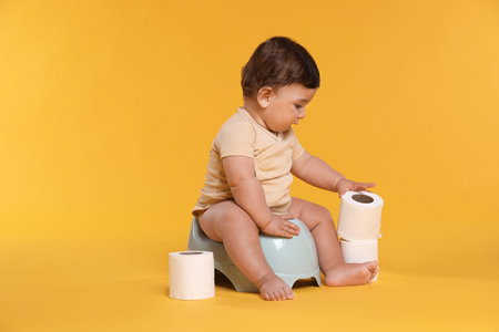 Cute little baby sitting on potty and rolls of toilet paper against orange background