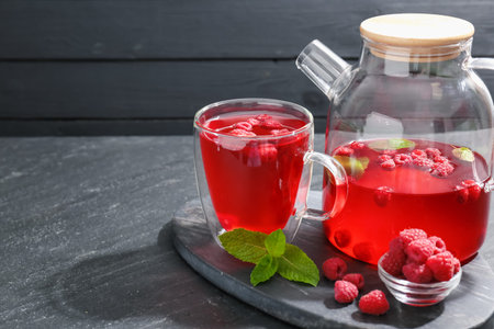Aromatic raspberry tea in glass cup, teapot, berries and mint on black table, closeup. Space for...