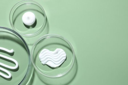 Different cosmetic products in Petri dishes on green background, flat lay. Space for text