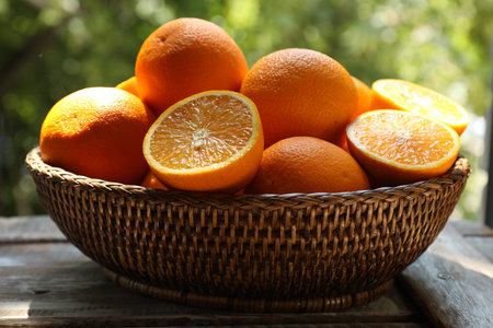 Fresh whole and cut oranges in wicker basket on wooden table against blurred green background,...