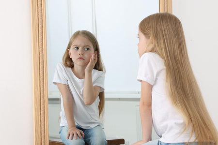 Upset little girl looking at herself in mirror indoorsの写真素材