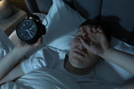 Frustrated man with alarm clock suffering from insomnia in bed indoors, above view