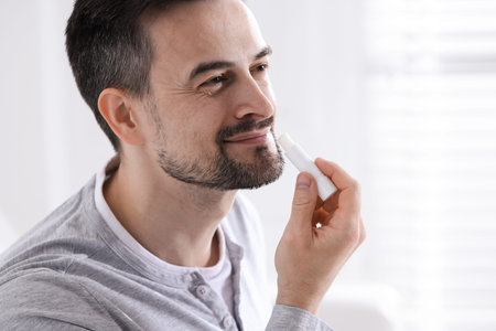 Handsome man applying lip balm onto his lips on blurred light background