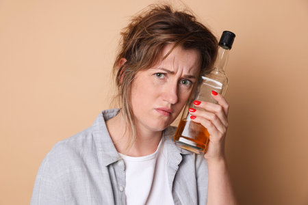 Woman with bottle of alcoholic drink suffering from hangover on beige backgroundの写真素材