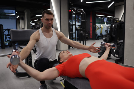 Young woman exercising with dumbbells with professional personal trainer in gymの写真素材