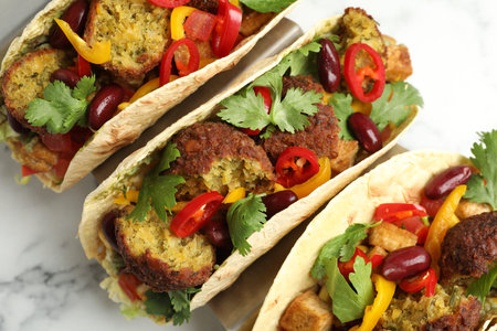 Tasty vegetarian tacos with falafel, tofu, beans and vegetables on white marble table, above view