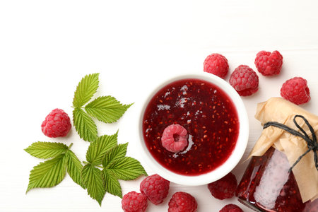 Sweet raspberry jam, berries and green leaves on white wooden table, flat lay