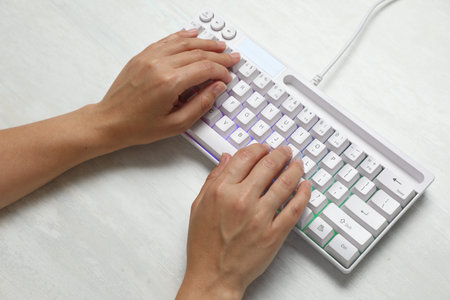 Woman typing on modern keyboard with RGB lighting at white wooden table, above view