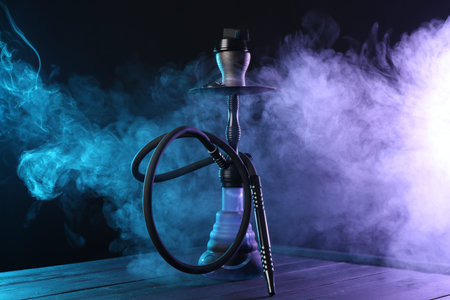 Hookah on wooden table with smoke in color lights against black background
