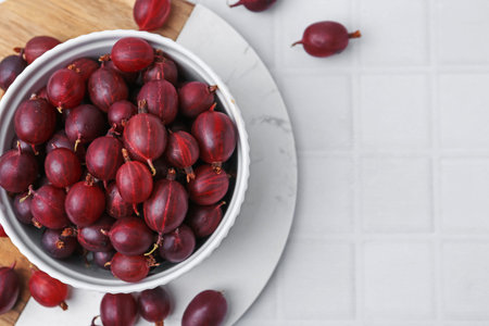 Ripe red gooseberries in bowl on white tiled table, flat lay. Space for text