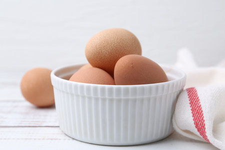 Raw chicken eggs in bowl on white wooden table, closeup