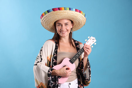 Happy woman in sombrero hat playing ukulele on light blue backgroundの写真素材