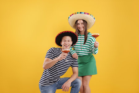 Happy couple in sombrero and Flamenco hats with cocktails on yellow backgroundの写真素材