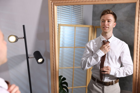 Handsome man adjusting necktie while getting ready near mirror indoorsの写真素材