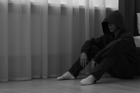Sadness and depression. Man in hoodie near curtains at home, space for text. Black and white effectの写真素材