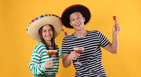 Happy couple in sombrero and Flamenco hats. Man and woman with cocktails and maracas on yellow...