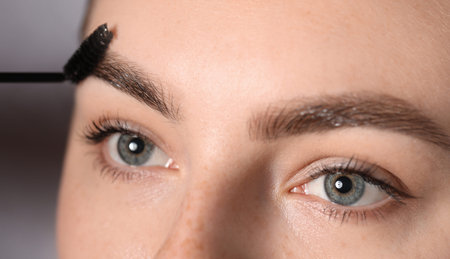 Young woman brushing her eyebrow with spoolie on light grey background, closeup