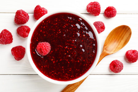 Sweet raspberry jam in bowl and berries on white wooden table, flat lay