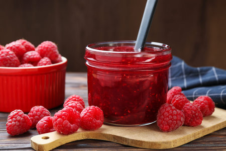 Sweet raspberry jam in glass jar and berries on table, closeup
