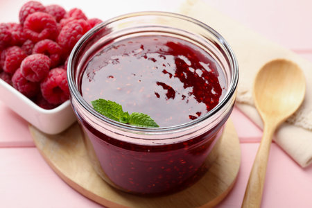 Sweet raspberry jam in glass jar, berries and mint on pink wooden table, closeup