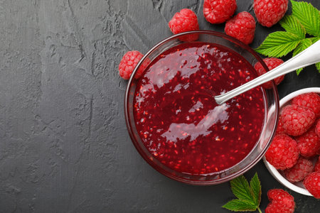 Sweet raspberry jam in glass bowl and berries on gray textured table, flat lay. Space for text