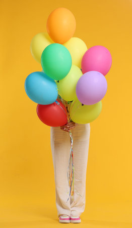 Woman hiding behind bunch of colorful balloons on yellow background