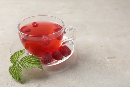 Tasty raspberry tea in glass cup, berries and green leaves on light grey table, closeup. Space for...