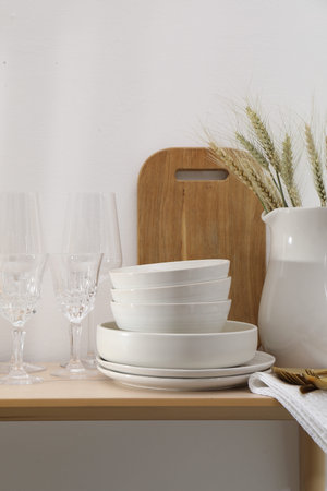 Different clean white dishware, cutlery, cutting board, glasses on wooden table indoors