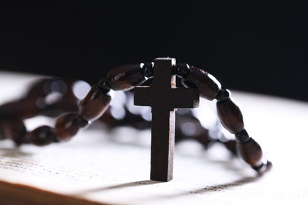 Rosary with wooden cross on Holy Bible against black background, closeup