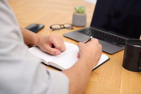 Copywriter with laptop writing in notebook at wooden table indoors, closeup
