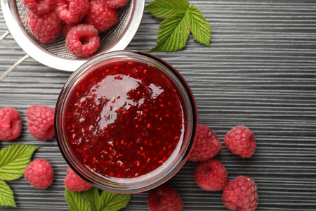 Sweet raspberry jam in glass jar and berries on dark gray wooden table, flat lay