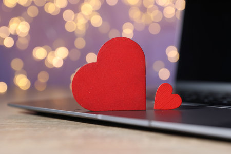 Online dating site. Red hearts and laptop on wooden table against blurred lights, closeup. Bokeh effectの写真素材