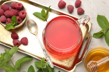 Tasty raspberry tea, berries, honey and mint on light grey table, flat lay