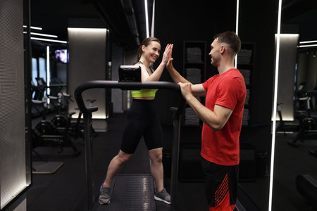 Young woman exercising on treadmill with professional personal trainer and giving high five in gym
