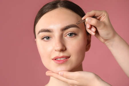 Cosmetologist plucking young woman's eyebrow with tweezers on pink background, closeup