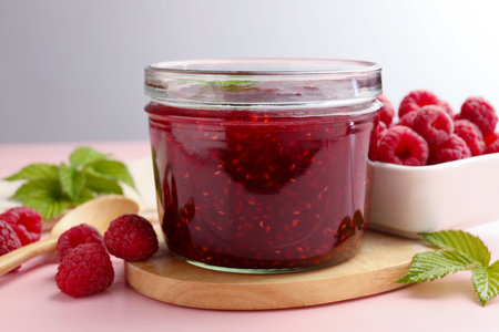 Sweet raspberry jam in glass jar, berries and mint on pink wooden table, closeup