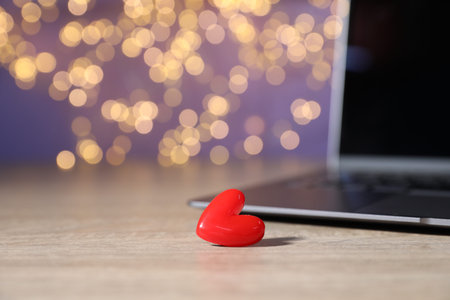 Online dating site. Red heart and laptop on wooden table against blurred lights, closeup. Bokeh...