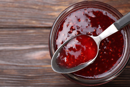 Sweet raspberry jam in glass jar and spoon on wooden table, top view. Space for text