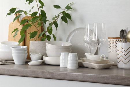 Different clean white dishware, wooden cutting boards, glasses, cutlery and houseplant on counter in...