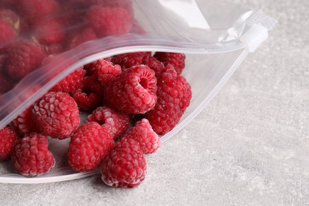 Ripe frozen raspberries in open zipper bag on gray textured table, closeup