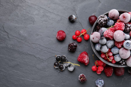 Mix of ripe frozen berries and bowl on dark textured table, flat lay. Space for text