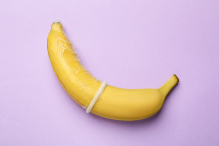 Sex education. Banana with condom on violet background, top view