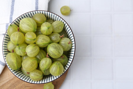 Fresh green gooseberries in bowl on white tiled table, top view. Space for textの写真素材