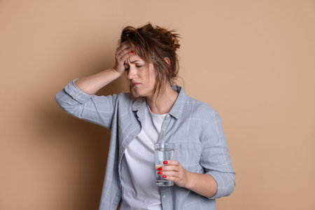 Woman with glass of water suffering from hangover on beige background